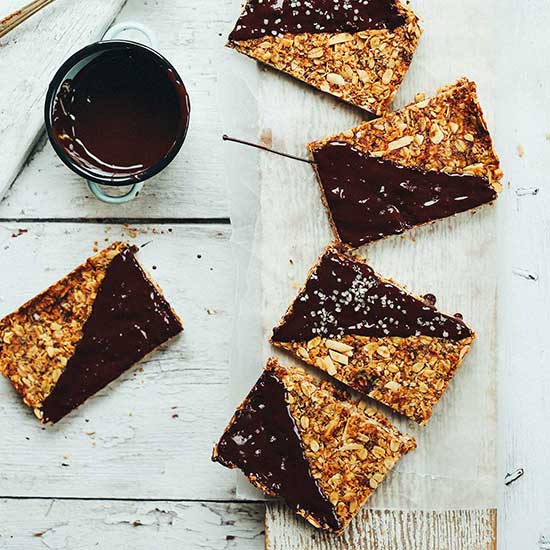 Melted chocolate next to several crunchy baked granola bars half dipped in chocolate