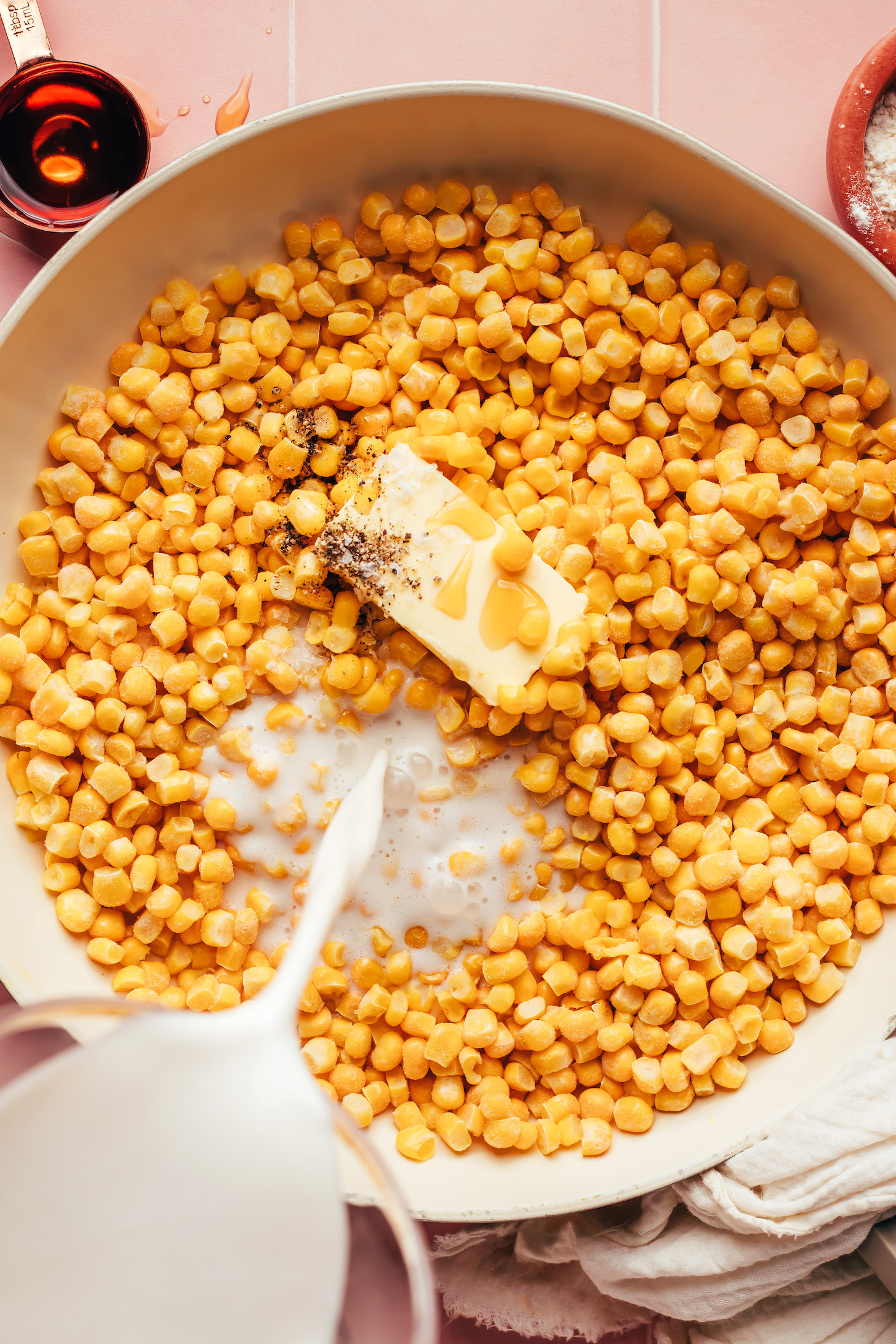 Pouring cashew milk over frozen corn, vegan butter, maple syrup, and black pepper