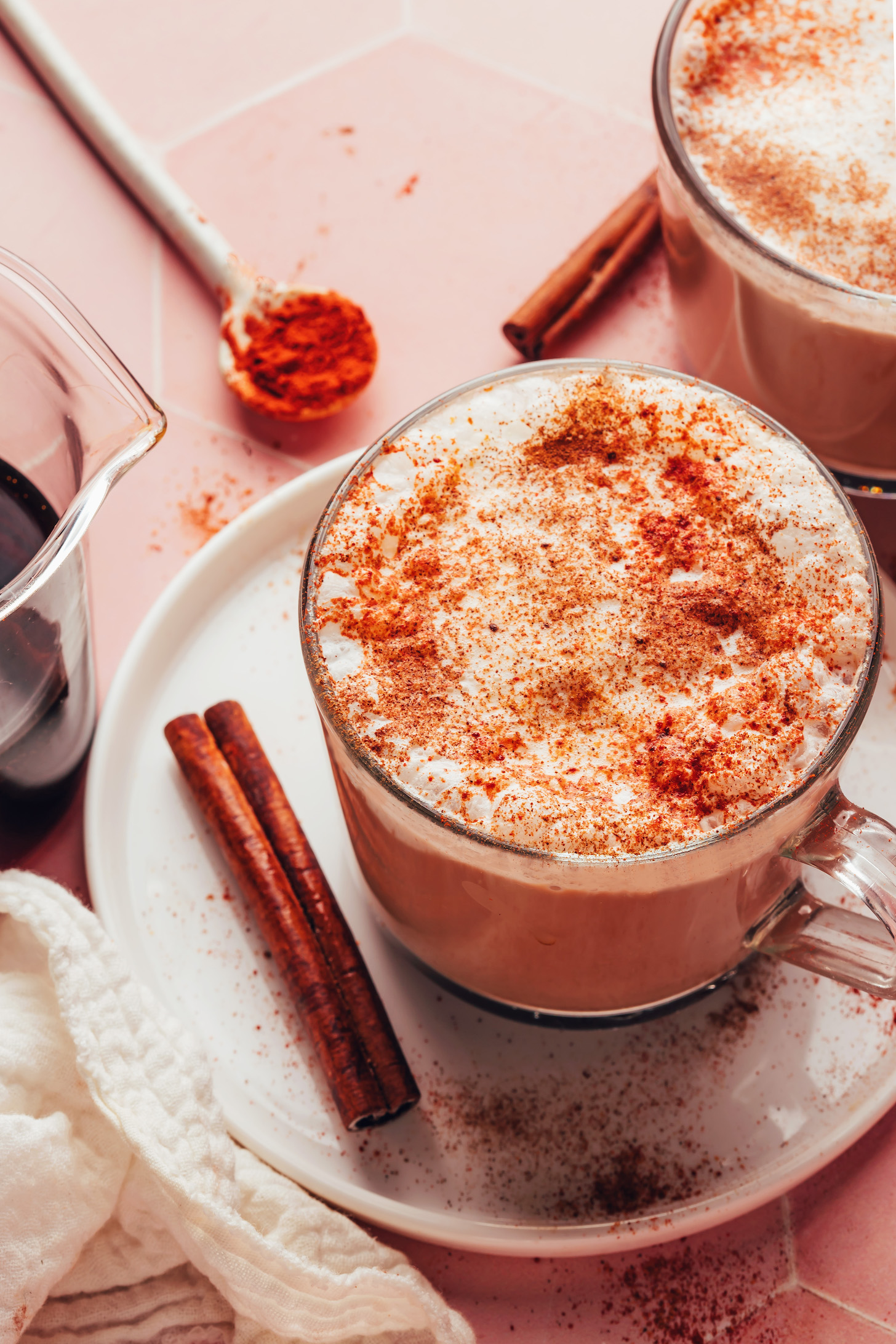 Cinnamon stick, cayenne, and maple syrup next to two mugs of our creamy Mayan mocha made with coffee or dandelion root